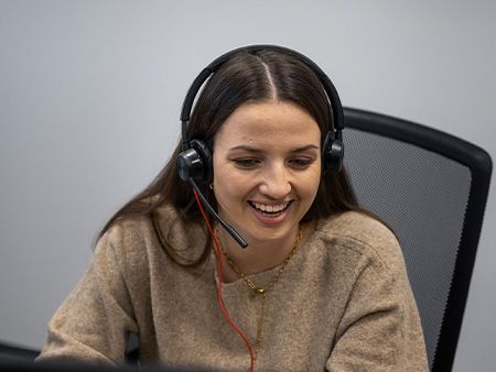 Eine Frau mit langen, dunklen Haaren trägt ein Headset mit Mikrofon und sitzt auf einem Bürostuhl. Sie lächelt freundlich, während sie konzentriert arbeitet. Sie trägt einen beigefarbenen Pullover und eine goldene Kette. Der Hintergrund ist einfarbig.