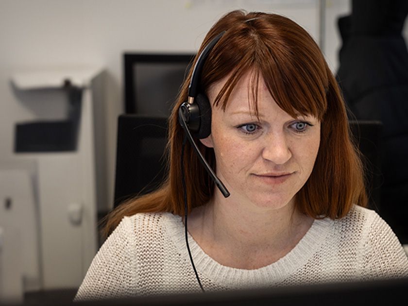 Eine Frau mit schulterlangem, roten Haar trägt ein Headset mit Mikrofon und blickt auf einen Bildschirm. Sie trägt einen weißen Strickpullover. Im Hintergrund sind unscharf Büromöbel und eine dunkle Jacke auf einem Stuhl zu erkennen.